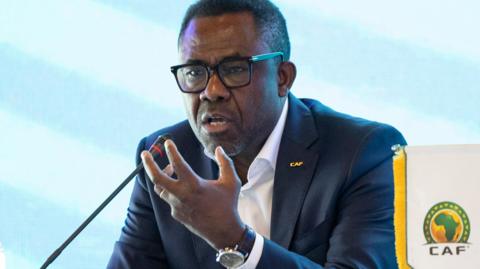 Veron Mosengo-Omba wearing a dark blue suit jacket and white shirt raises his left hand and speaks into a microphone at a press conference while a pendant displaying the green and yello Caf log sits to the side of him