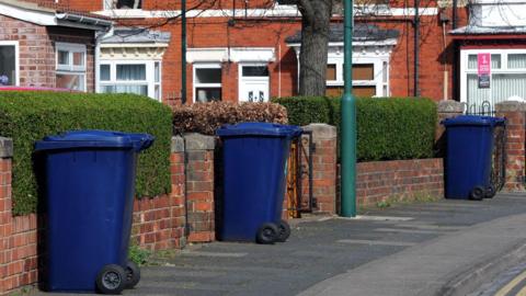 Three blue bins line up outside terraced houses. 