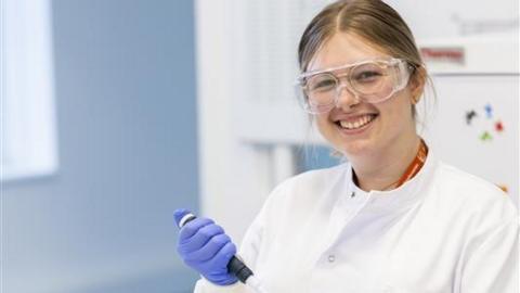 Millie-Rose Doolan in a white lab coat and blue gloves. She has dark blonde hair that has been tied back and is also wearing protective glasses. 