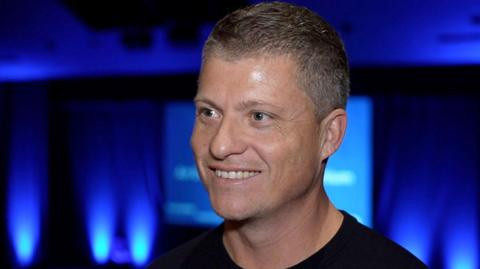 Justin Rees has short grey/black hair and is wearing a black T-shirt. He is smiling in front of what appears to be a stage illuminated by blue lights.