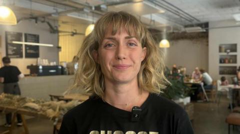 Anna Wiltshire looks at the camera. She has thick, shoulder-length, blonde hair and is wearing  a black t-shirt. Behind her are people sat at various wooden tables in the cafe. 