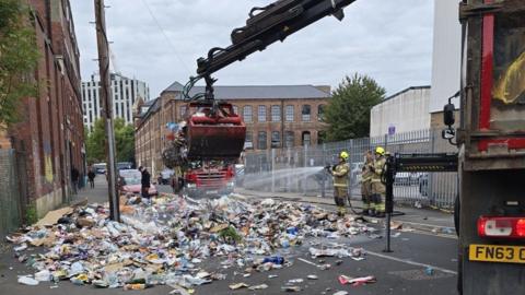 A city street blocked by a pile of paper and plastic, which is being sprayed with water as a crane with a grabber arm picks up quantities of the material.