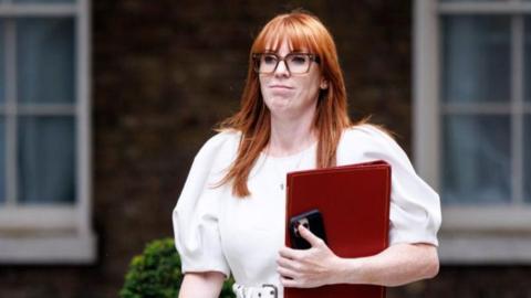 Rayner walks along, holding a red file and wearing a white dress.