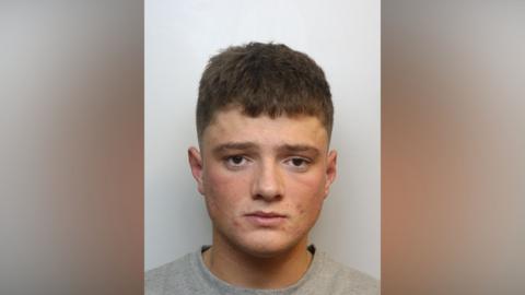 A mugshot of a young white man with brown hair and spots on his face, wearing a grey jumper.