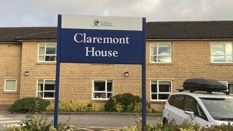 Sign saying 'Claremont House' in forefront of picture, with a two storey sandy brick building in the background. There is a white car parked in the car park to the right of the picture. 