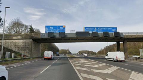 The M25 at junction 12, with blue signs over the top of the carriageway showing the way to Basingstoke, Southampton and Richmond on the M3 and towards Heathrow and Watford on the M25. The road is divided by a white chevroned section and there are some cars and vans on the road.
