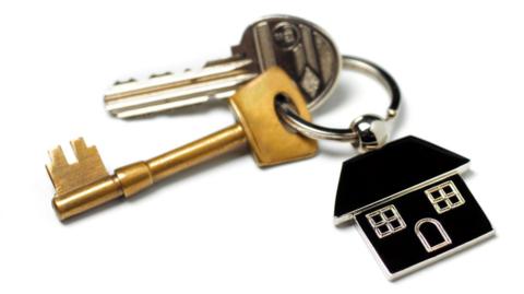 A set of house keys - one silver-coloured and the other gold-coloured - attached to a black key ring in the shape of a house. The keys are on a white background.