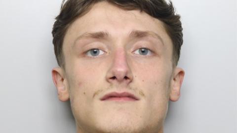 A mugshot of a 20-year-old man with blue eyes, pale skin, brown pushed back hair and a thin patchy moustache. 