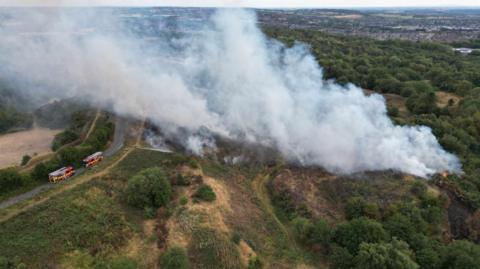 An aerial photograph of a large wildfire on an area of grassland. There are two fire engines in the bottom left hand corner of the image. There is a huge plume of smoke stretching from the right hand side of the image across the park.
