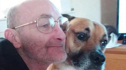 Henry White wearing a black top and glasses. He is hugging a brown and black dog in a bedroom.