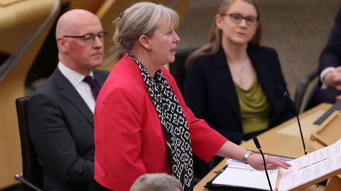 Shona Robison, wearing a red jacket, stands in the Scottish Parliament to deliver her Budget speech
