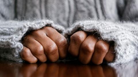 A stock image of hands in fists, resting on a wooden surface. The person is wearing a grey woolly jumper and its sleeves are pulled up close to the person's knuckles.