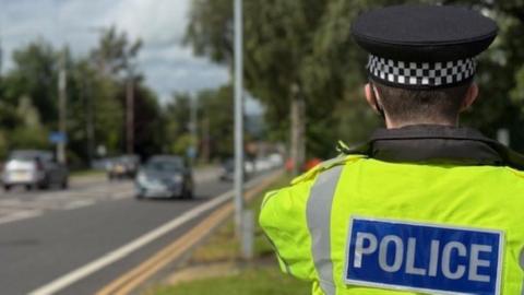 A picture of a police officer standing with his back towards the camera on a road. He is wearing a police hat and high-vis jacket with the word: Police on the back.
