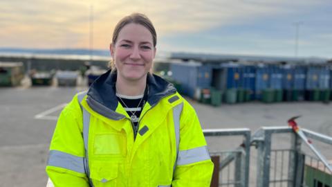 Sarah Robinson, Operations Manager, stood in a yellow high viz coat and a striped black and white jumper is poking out underneath. Her light brown hair is tied back and she is smiling. She is stood on a balcony at the Longue Houge waste site, where recycling bins are in the blurred background of the image. The sky is a dusk light, with peach clouds and darkening blue.  