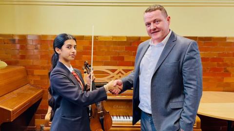 A teenage girl in school uniform holds a violin and bow while shaking hands with a man in a grey blazer and open-collared shirt. They are standing in front of an upright Cramer piano in a room with glazed brown brick walls and wooden flooring. The setting appears to be a traditional music school or rehearsal hall.
