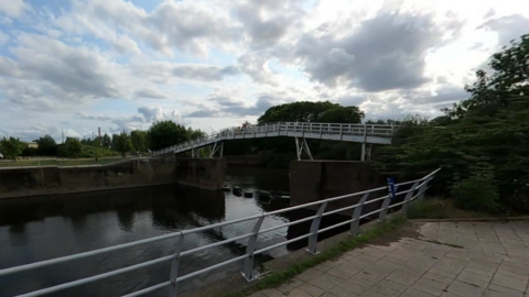 A small footbridge pictured going across a square body of water. The bridge is narrow and grey in colour. There is a large tree at one side of the bridge. 
