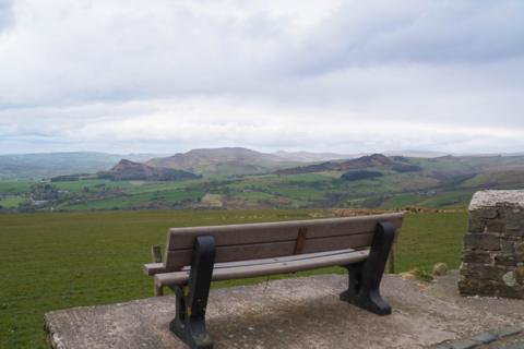 A wooden bench overlooking a wide, green countryside of rolling hills and patchwork fields under a cloudy, overcast sky, with a stone marker beside it.