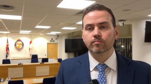 A man in his 30s with short hair and a trimmed beard looking towards the camera. He's wearing a blue suit and tie with a white shirt. Behind him are desks and chairs in a council building. At the back out of focus is a coat of arms with the union jack flag on the left and St George's flag on the right.