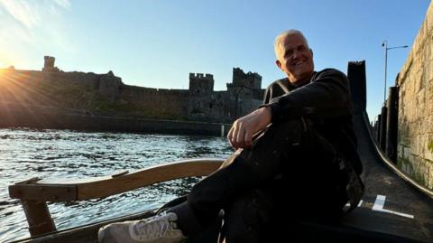 Nigel Rawlinson is sitting in a Viking-style longboat in Peel harbour on a sunny day with cloudless blue skies. Peel Castle is largely in silhouette behind him as the low sun sets.