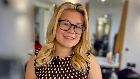 Jessica Flaxman stands in a hair salon with the background blurred out. She has long blonde hair and wears black-rimmed glasses. She is dressed in a brown dress with white polka dots.