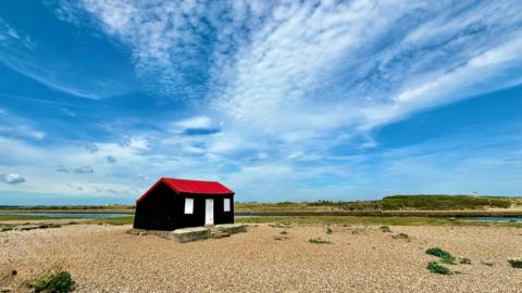 A black cabin with a red roof on the coast, on a pebble beach beneath a bright blue sky
