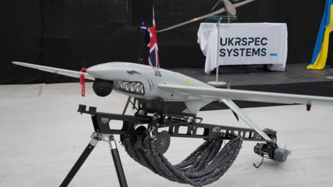 A Ukrspecsystems drone with a shark face and design on its nose propped up on a black metal stand. In the background there is a white banner which reads Ukrspecsystems and a Ukraine flag.