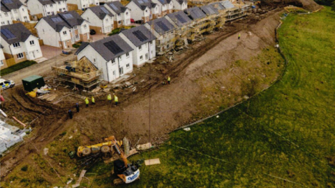 Ariel view of a housing construction site with rows of new-build houses - some are complete, some have scaffolding. A large construction vehicle lies at the bottom of a mud slope on grass.