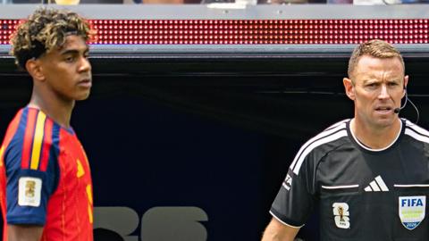 A close up photo of Stuart Burt in a black referee top with an earpiece and microphone. On the other side of the picture is Spanish forward Lamine Yamal in a red Spanish football shirt. Both are looking at the action off camera.