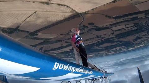 A woman is strapped to the wing of a plane which is flying upside down over brown fields.