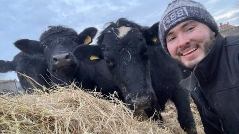 Harry Churchill in a selfie, on the right hand side of the picture and smiling as he leans into a black cow which is looking at the camera and leaning on another black cow that is also looking at the camera. There is straw in the foreground of the picture and you can see the blue sky behind. 