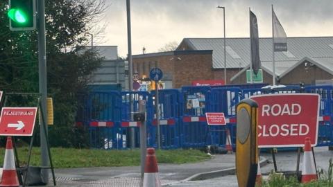 Junction into road with blue barriers across and many red and white road signs and cones.