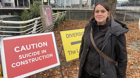 Sharon Wall smiling in front of the official-looking road signs which say Caution Suicide in Construction in red. And a yellow sign that says Diversion The Lost City. Sharon is wearing a black jumper and puffa jacket and has her hair tied up. 