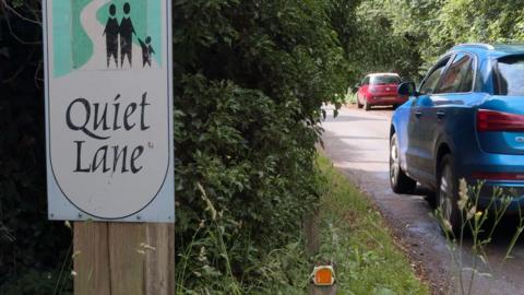 A roadside sign with the words "Quiet Lane" printed on it. To the right there is a road with a passing blue car.