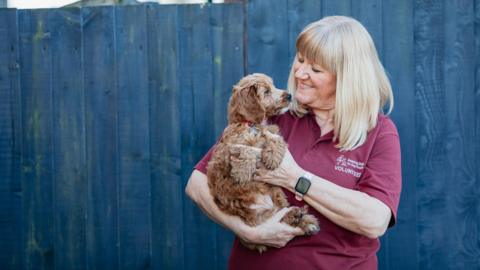 A blonde woman in a maroon t-shirt holding a chestnut-coloured puppy. 