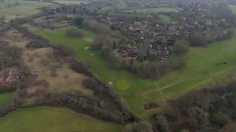 An aerial photography shows the area of the Abbey golf course that would be developed, as well as partially-wooded scrubland to the west. The existing Hither Green Lane estate is visible.
