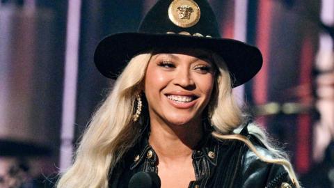 ǲԳé on stage smiling as she receives an award. She's styled in a black cowboy hat with a gold medallion on the front and a black denim jacket. She has long platinum blonde hair and wears large gold hoop earrings. 