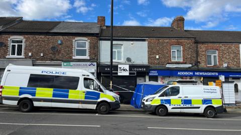 Two white police vans parked outside a row of shops. There is a blue police tent on the pavement and a police cordon.