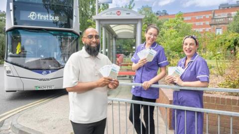 A First Bus representative and two female maternity staff stood on a pavement by a road. They are each holding travel tickets in their hands. Behind them is a parked double decker bus which reads 'First bus' on a digital screen.