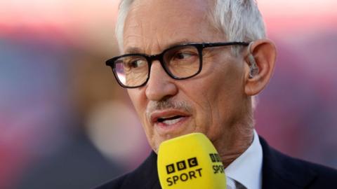 Gary Lineker speaking into a yellow BBC Sport microphone and looking off camera