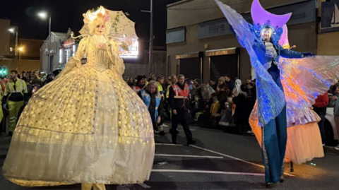Show a man and woman on stilts in brightly-coloured costumes with people in the background
