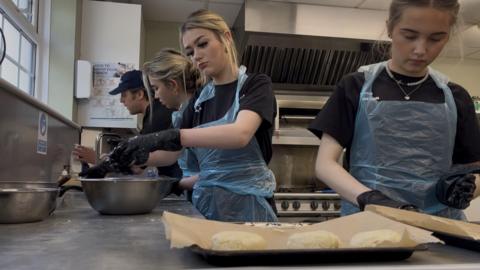 A man and three teenage girls in a kitchen standing over a work bench making scones.

The three girls with blonde hair are wearing black t-shirts and blue plastic aprons, 

There is a baking tray with dough in the shape of scones in the foreground