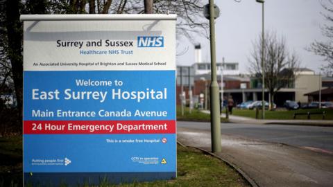 A large blue and white sign at the entrance to East Surrey Hospital shows directions to the main entrance and 24‑hour emergency department. The sign stands beside a road with streetlights, trees and hospital buildings visible in the background.