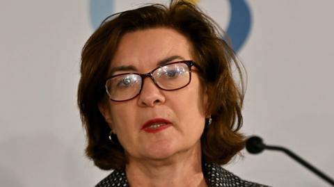 A head and shoulders picture of Eluned Morgan speaking into a microphone. She is wearing glasses, and a pokadot jacket. 