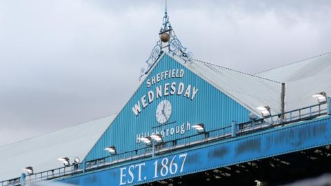 A sign above one of the signs at Hillsborough reads: 'Sheffield Wednesday; Hillsborough; Est. 1867' with a clock also on the stand 