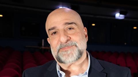 Comedian and actor Omid Djalili smiles at the camera while standing in the Ipswich Regent Theatre. He is bald and has a grey goatee beard. He is wearing a navy suit jacket with a blue shirt. Red seats of the theatre can be seen behind him.