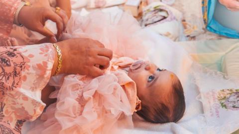 Sophia Kelemen is lying on her back looking up at an adult whose hands can be seen on her. She is wearing a frilly pink dress and is lying on a blanket monogrammed with the name Sophia Beatrice. Sophia is sucking on a baby pink dummy and has bright blue eyes 