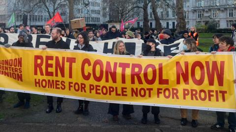 A group of protesters gathered outdoors, holding a large yellow banner with red text that reads, "rent control now - homes for people not for profit," alongside the "London Renters Union" logo. There are additional banners and signs visible in the background, with a crowd of individuals standing in a park.