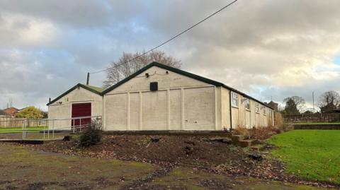Two single-storey, white-walled buildings with dark rooves and a ramp leading to the entrance, with a muddy car park in front