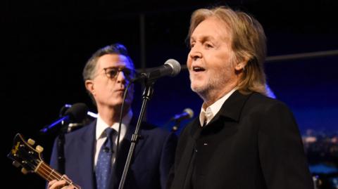 Paul McCartney playing guitar with Stephen Colbert behind him