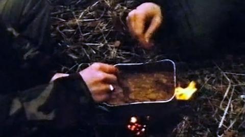 Hand of a soldier stirring a portion of food being cooked over a campfire outdoors.